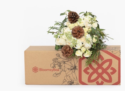 Elegant floral arrangement with white flowers and green foliage atop a BloomsyBox cardboard box, displaying a distinctive flower logo and pattern on a neutral background.