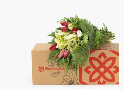 A vibrant bouquet of red tulips and white flowers with lush greenery, partially wrapped and emerging from a BloomsBox branded cardboard box on a white background.