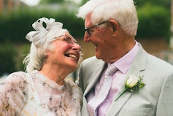 Elderly couple smiling and embracing in celebration, with the man in a light suit and the woman wearing a floral dress and fascinator, conveying joy and affection.