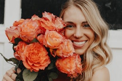 Smiling woman with blonde hair holding a large bouquet of orange roses close to her face, standing in front of a white background.
