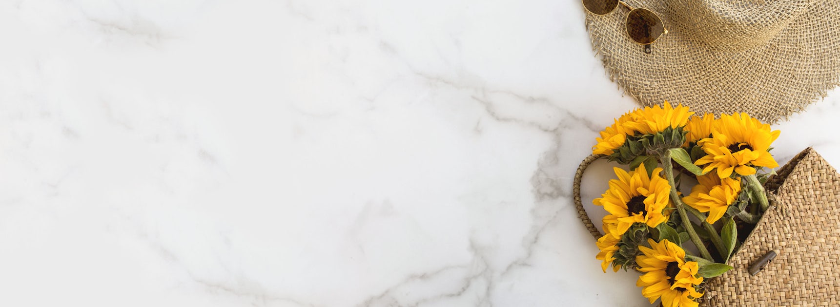 Sunflowers nestled in a woven tote alongside a stylish sun hat and sunglasses on marble.