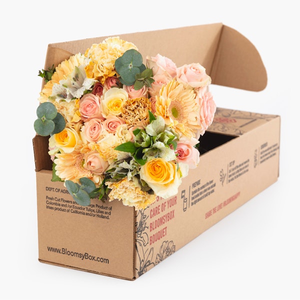 A vibrant bouquet of yellow and pink roses with greenery, beautifully arranged and ready to be unpacked from a BloomysBox cardboard delivery box.