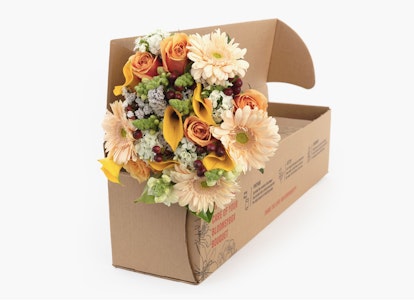 A vibrant bouquet of fresh flowers with roses, sunflowers, and daisies arranged in a cardboard delivery box against a white background, symbolizing a special occasion gift.