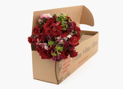 A vibrant bouquet of red and pink flowers, including roses and peonies, arranged in a brown cardboard delivery box against a white backdrop.