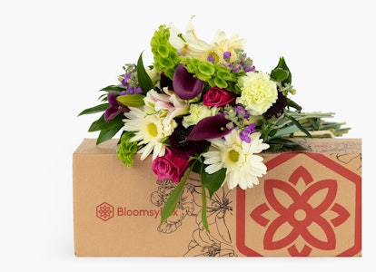 Beautiful bouquet of flowers with lilies and roses beside a cardboard delivery box with a flower logo, presented against a clean white background.