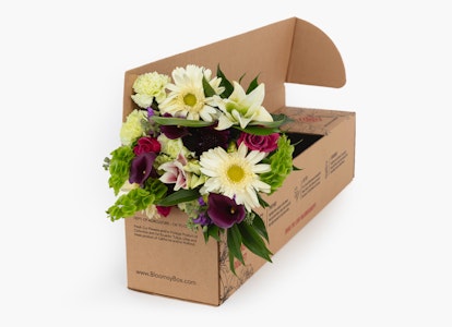 Bouquet of fresh flowers with white lilies, purple flowers, and green foliage packaged in an open cardboard box isolated on a white background.