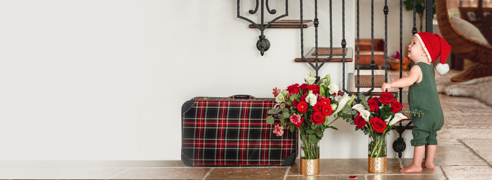 A charming toddler in a Santa hat admires a festive floral arrangement beside a vintage suitcase.