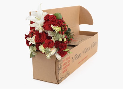 Elegant bouquet of red roses and white lilies with lush greenery, partially arranged inside an open cardboard box, against a clean white background.