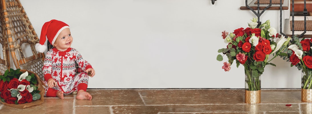 A joyful baby in a festive hat sits beside vibrant holiday flower arrangements.