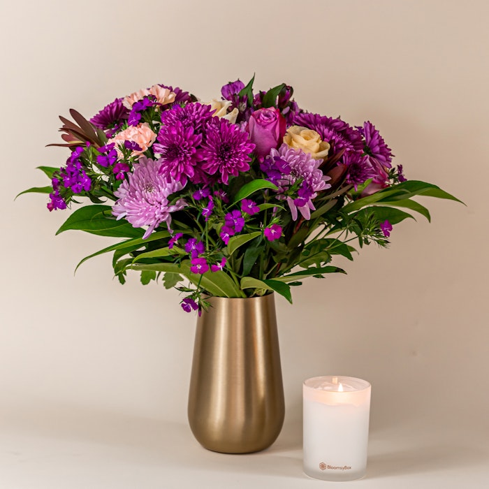 A vibrant bouquet of purple and pink flowers, including roses and chrysanthemums, arranged in a gold vase next to a lit scented candle on a neutral background.