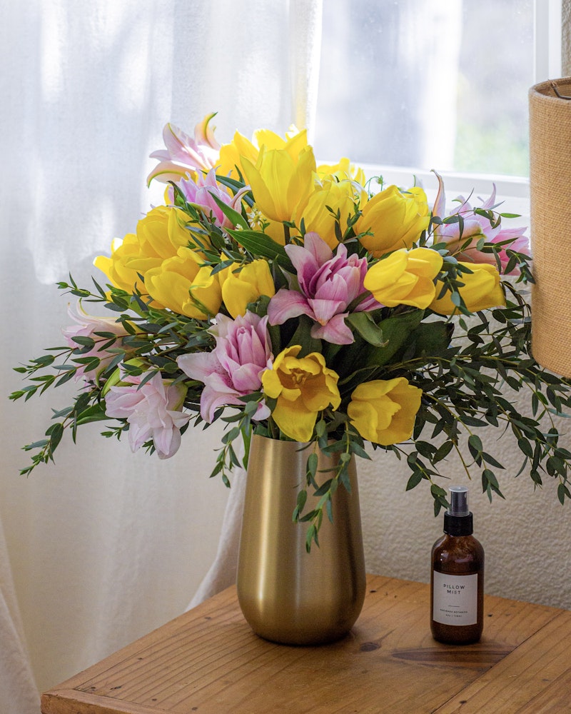 Vibrant yellow and pink flowers arranged in a golden vase on a wooden table, with translucent curtains and a brown bottle beside, creating a cozy, bright interior ambiance.