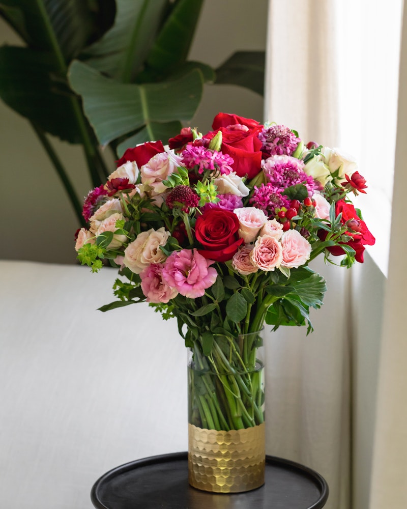 Elegant bouquet of fresh flowers including red roses, pink blooms, and purple flowers in a gold textured vase on a black tray by a window with greenery in the background.