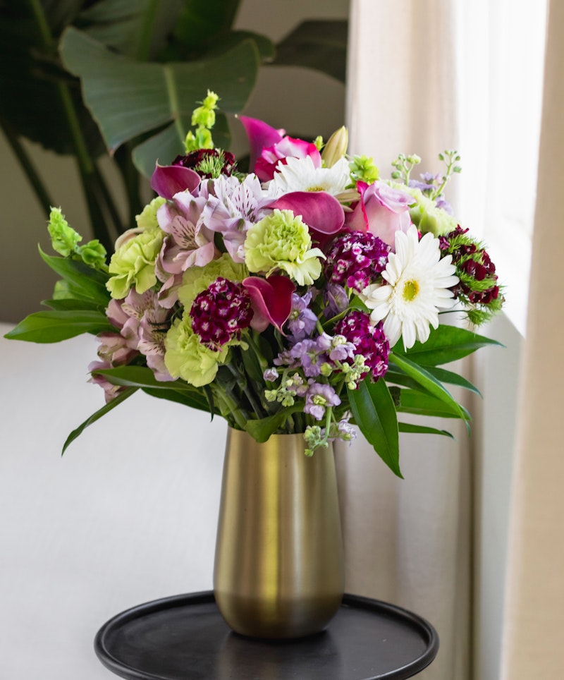 Vibrant bouquet of mixed flowers including purple orchids, pink lilies, white daisies, and greenery in a gold vase on a black stand by a window with sheer curtains.