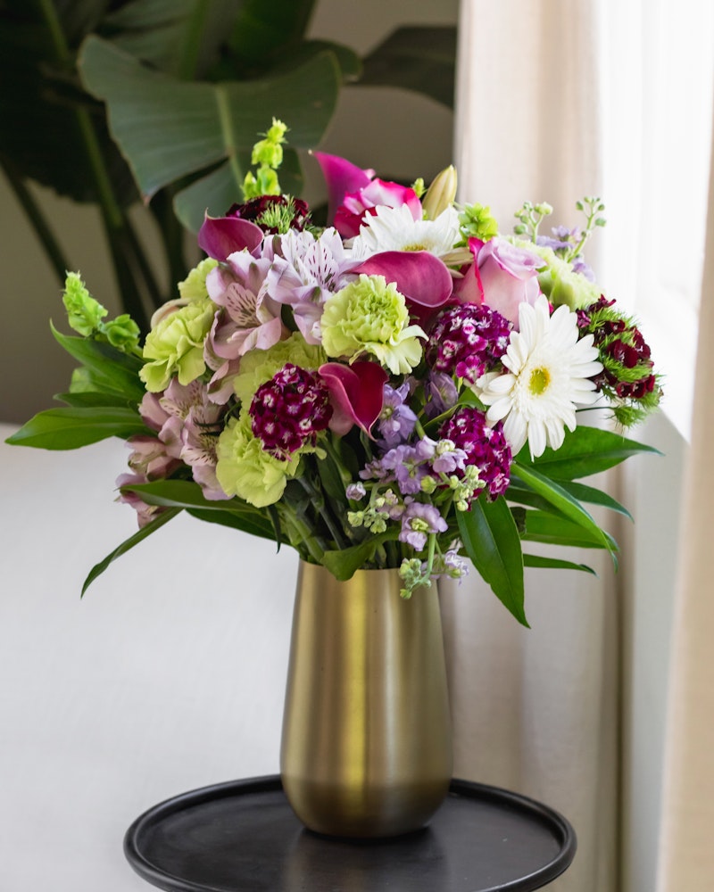 Vibrant bouquet of mixed flowers including purple orchids, pink lilies, white daisies, and greenery in a gold vase on a black stand by a window with sheer curtains.