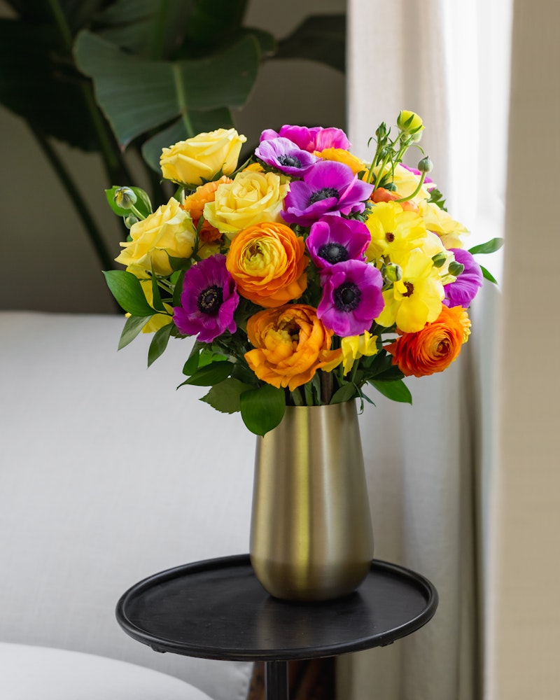 Vibrant bouquet of yellow, orange, and purple flowers in a golden vase on a black side table, with natural light filtering through a window.