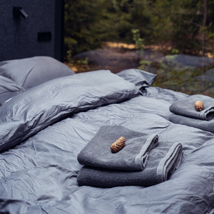 Cozy camp bedding setup with grey sleeping bags and pillows in a tent, facing a forest environment, featuring neatly placed towels and a pinecone on top.