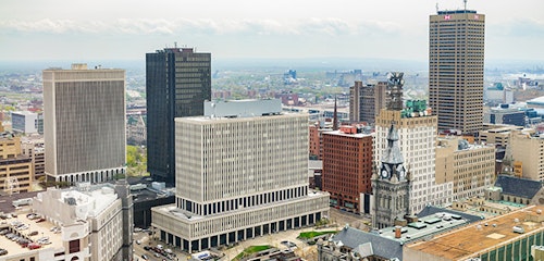 Aerial view of a bustling cityscape with a mix of modern high-rise buildings and historic architecture under a cloudy sky, showcasing the urban density and skyline.