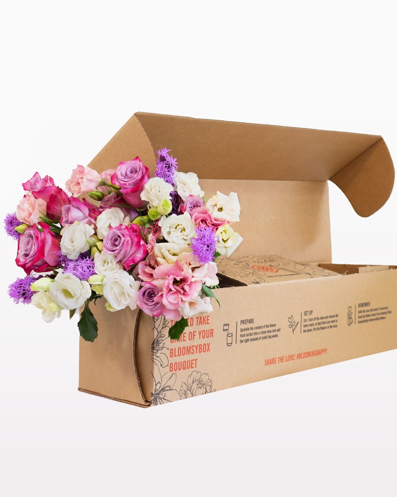 Colorful bouquet of pink and white roses with assorted flowers peeping out of a brown cardboard Bloomsbox, suggesting floral delivery or gift.