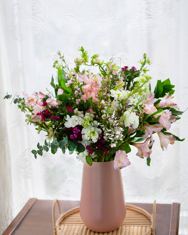 A vibrant bouquet of mixed flowers, including pink lilies and white blooms, arranged in a peach vase on a wooden tray against a white lace curtain backdrop.