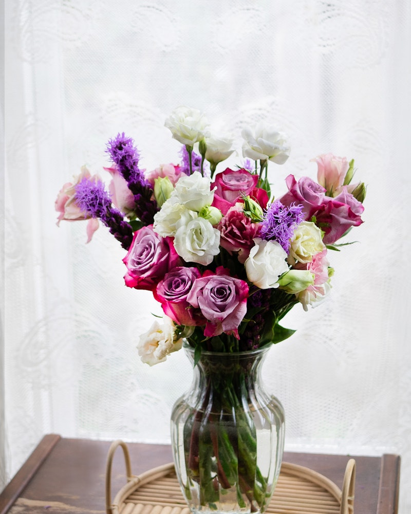 A vibrant bouquet of mixed flowers including purple, pink, and white blooms arranged in a clear glass vase on a wooden tray, with soft natural light filtering through a lace curtain.