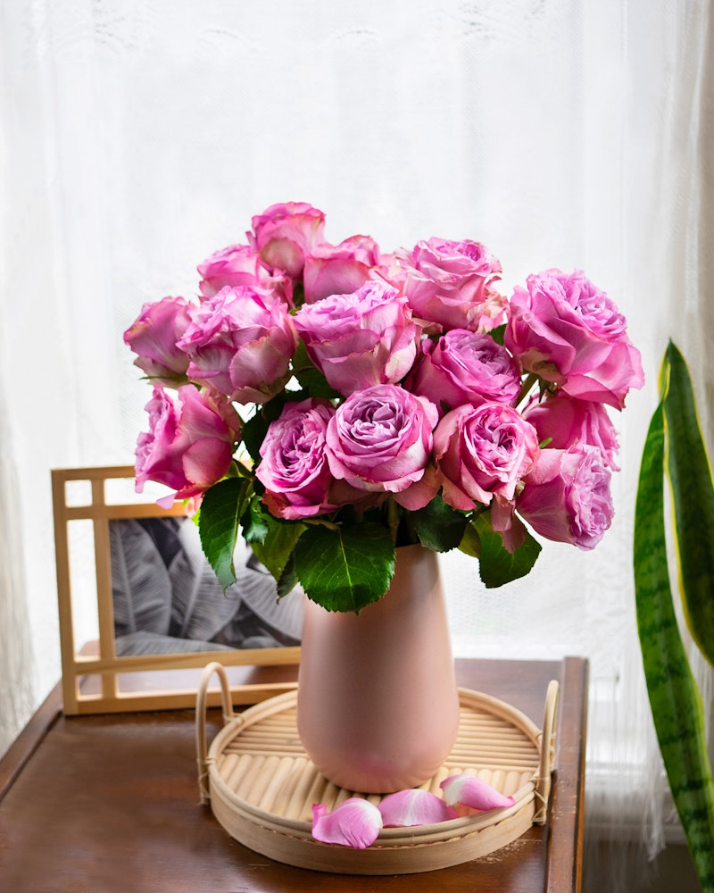 Vibrant bouquet of pink roses in a soft pink vase on a wooden tray beside a window with sheer curtains, with a few petals scattered around.