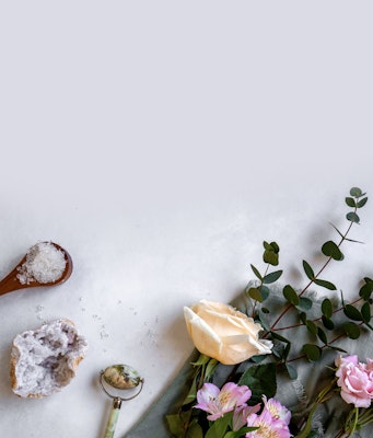 Flat lay of a pale background with spa essentials, including a scoop of sea salt, fresh flowers, and eucalyptus branches, arranged in a serene and aesthetic composition.