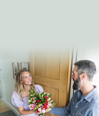 Man in a denim shirt presenting a bouquet of colorful flowers to a delighted woman with long blonde hair in a lilac top, indoors with a wooden door background.