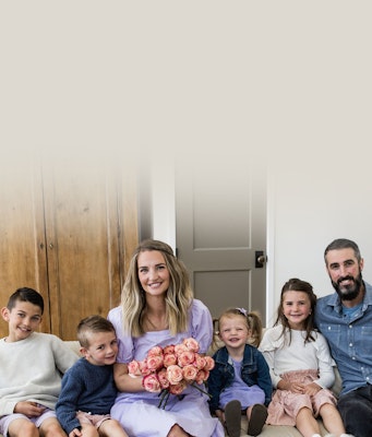 Family portrait of a smiling woman holding a bouquet of pink roses, seated with four children and a bearded man against a neutral background with a wooden door.