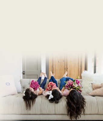 Three people lounging on a couch with their feet up on the backrest, surrounded by fluffy pillows and each holding a bright bouquet of flowers, in a cozy home setting.