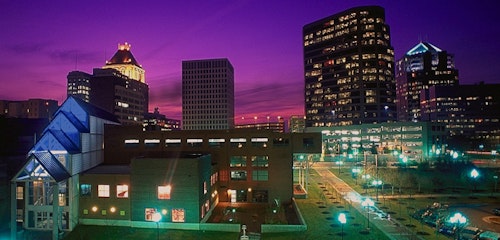 Twilight cityscape showing illuminated buildings with a striking purple sky, highlighting architectural diversity from modern glass structures to traditional skyscrapers.