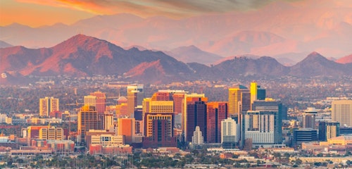 Sunset view of a bustling city skyline with prominent high-rise buildings against a backdrop of distant mountains under a golden orange sky.