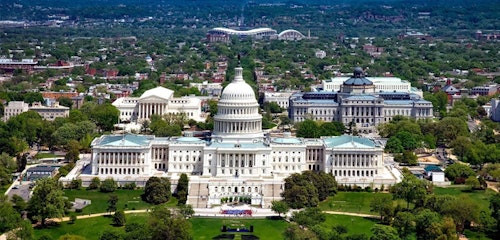 Aerial view of the United States Capitol Building with lush greenery, surrounding streets, and distant Washington D.C. landmarks under a clear sky.
