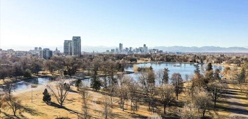 A panoramic view of a city skyline with tall buildings, a large park with trees and pathways, and a tranquil lake on a bright, sunny day with clear skies.