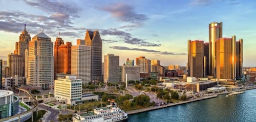 Panoramic view of a city skyline at sunset featuring prominent high-rise buildings, with the sunlight reflecting off, adjacent to a calm river with a boat.