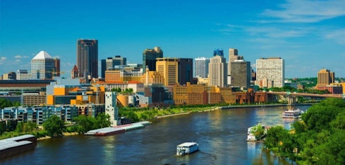 Scenic view of a vibrant city skyline with modern buildings beside a tranquil river, featuring passing boats and lush greenery under a clear blue sky.
