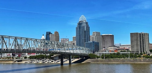 Panoramic view of a cityscape with a distinctive skyscraper and a truss bridge crossing a river, under a clear blue sky with sparse clouds. Urban skyline with water reflections.