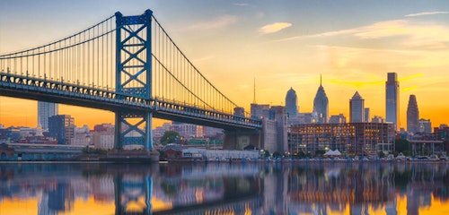 Sunset view of Philadelphia skyline with the Benjamin Franklin Bridge in the forefront, reflecting on the calm waters below, against a vibrant orange sky.