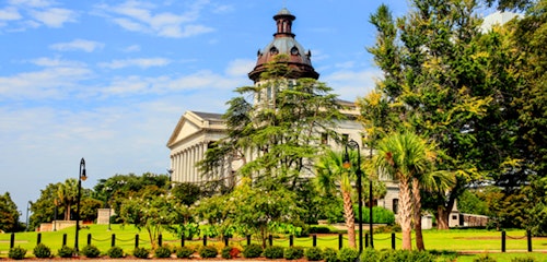 Historic white building with a central dome surrounded by lush greenery and tall palm trees under a clear blue sky, illustrating classic architecture in a serene park setting.