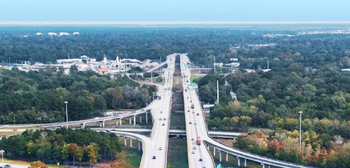 Elevated view of a busy highway with multiple overpasses and lanes, surrounded by greenery with a clear sky above, showcasing infrastructure and transportation.