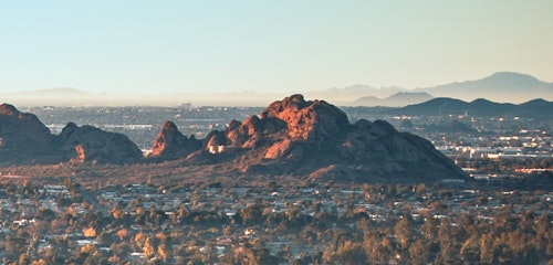 Scenic view of a cityscape at dawn with soft light hitting the rocky hills in the foreground and a hazy skyline in the distance under a pale blue sky.