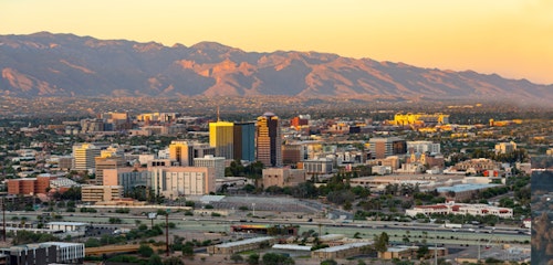 Golden hour view of a bustling city skyline with modern buildings against a backdrop of distant mountains under a soft, pastel-colored sky.