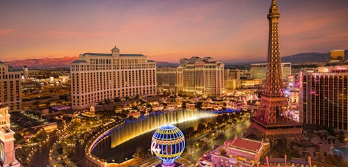 Aerial view of the Las Vegas Strip at dusk, featuring the Bellagio fountains, Paris Las Vegas's Eiffel Tower replica, and illuminated city landmarks under a colorful sky.