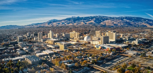 Aerial view of a sprawling cityscape with high-rise buildings under a clear blue sky, with a backdrop of large mountain range and patches of greenery.