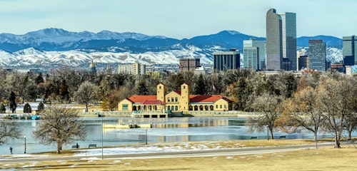 Panoramic view of a city skyline with modern buildings against a backdrop of distant snow-capped mountains, with a park and partially frozen lake in the foreground.