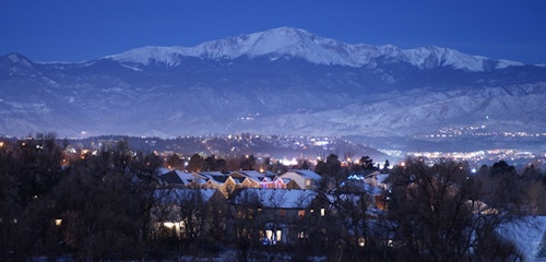 Twilight descends on a snow-capped mountain towering over a cityscape with illuminated buildings, highlighting the serene interaction between urban life and natural landscape.