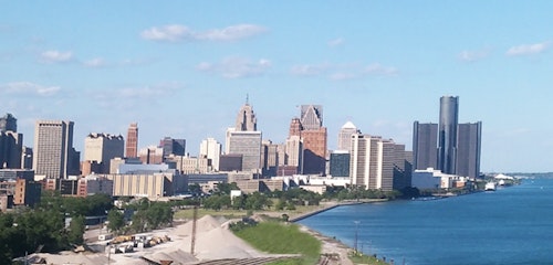 A panoramic view of a city skyline featuring tall skyscrapers under blue skies beside a large river, highlighted by sunshine with a clear view of waterfront buildings.