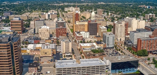 Aerial view of a bustling downtown district with a mix of modern and historic buildings, busy streets, and lush greenery on the outskirts under a clear sky.