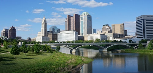 Panoramic view of a city skyline featuring modern high-rise buildings, a bridge crossing a calm river, and lush greenery under a clear blue sky with fluffy clouds.