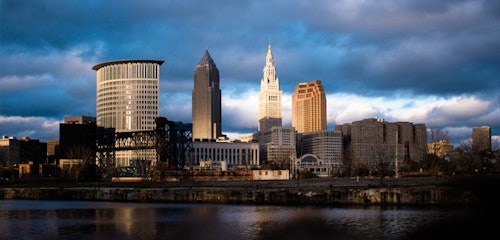 Dramatic skyline view of Cleveland with prominent buildings reflected in the water under a cloudy sky with a glimpse of sunlight illuminating the cityscape.