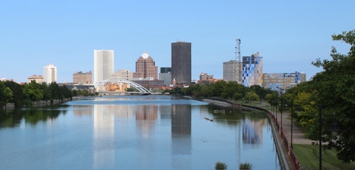 Scenic view of a city skyline with modern buildings reflected in a calm river, a clear blue sky overhead, and green trees lining the waterfront.